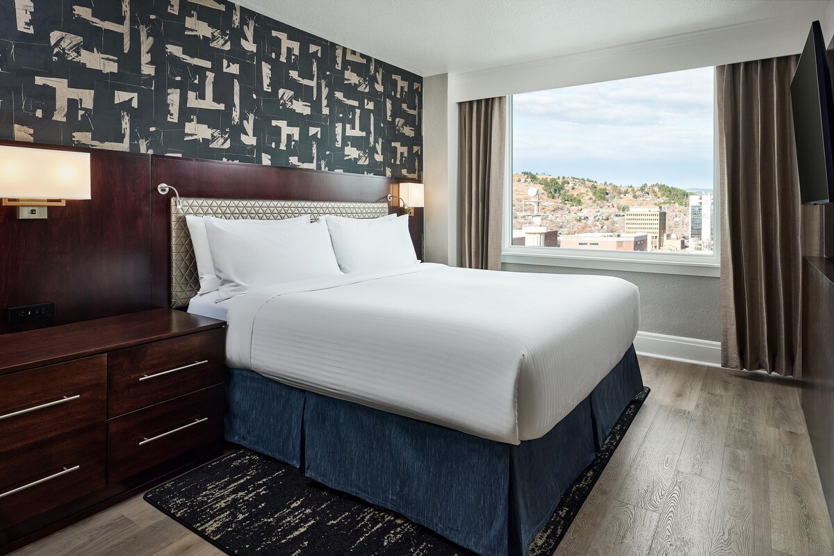 Quarto de hotel com uma cama grande, uma cômoda de madeira, uma mesa de cabeceira com abajur e vista para a cidade.