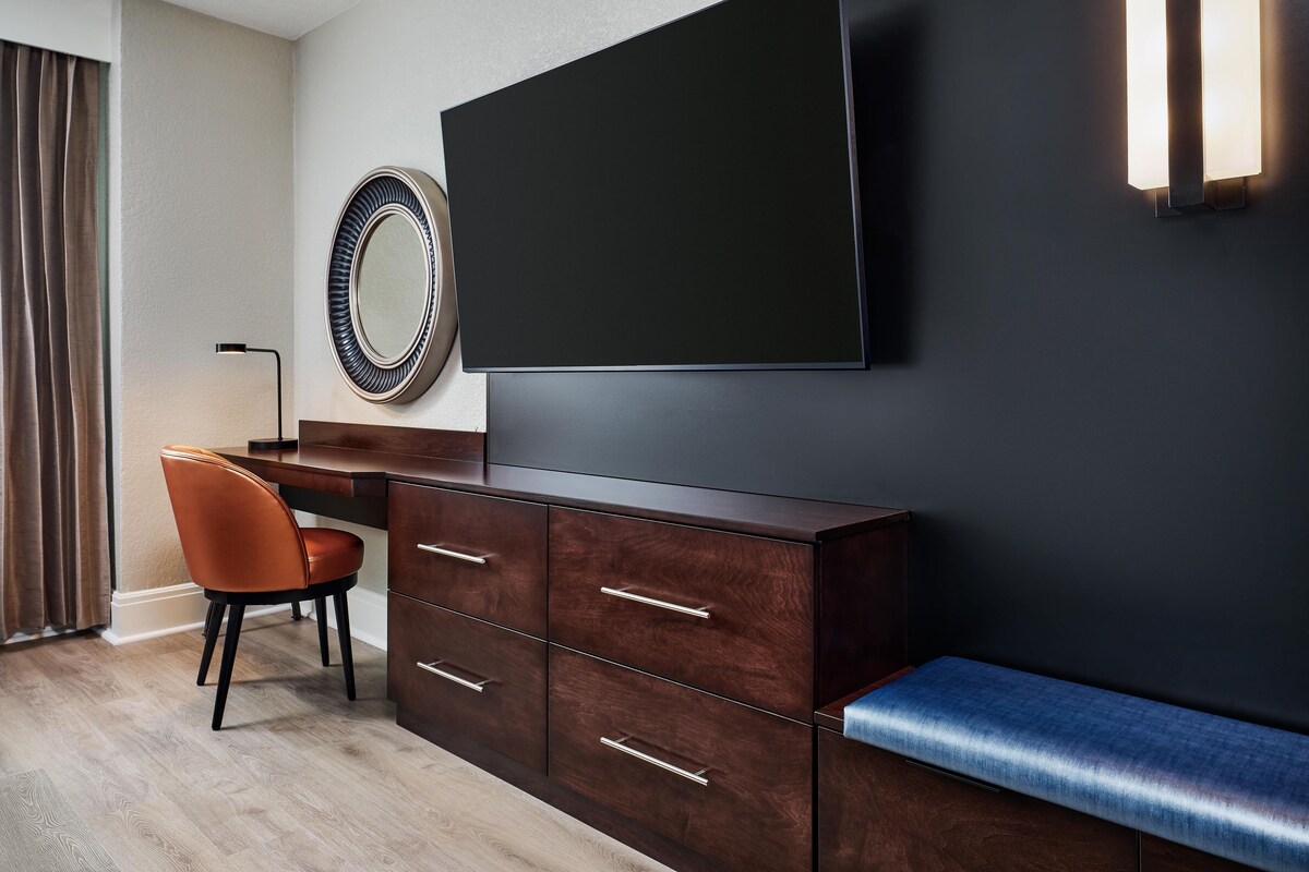Quarto moderno com parede escura, uma televisão de tela plana grande, um armário de madeira com gavetas, um espelho redondo, uma poltrona de couro marrom e um banco azul.