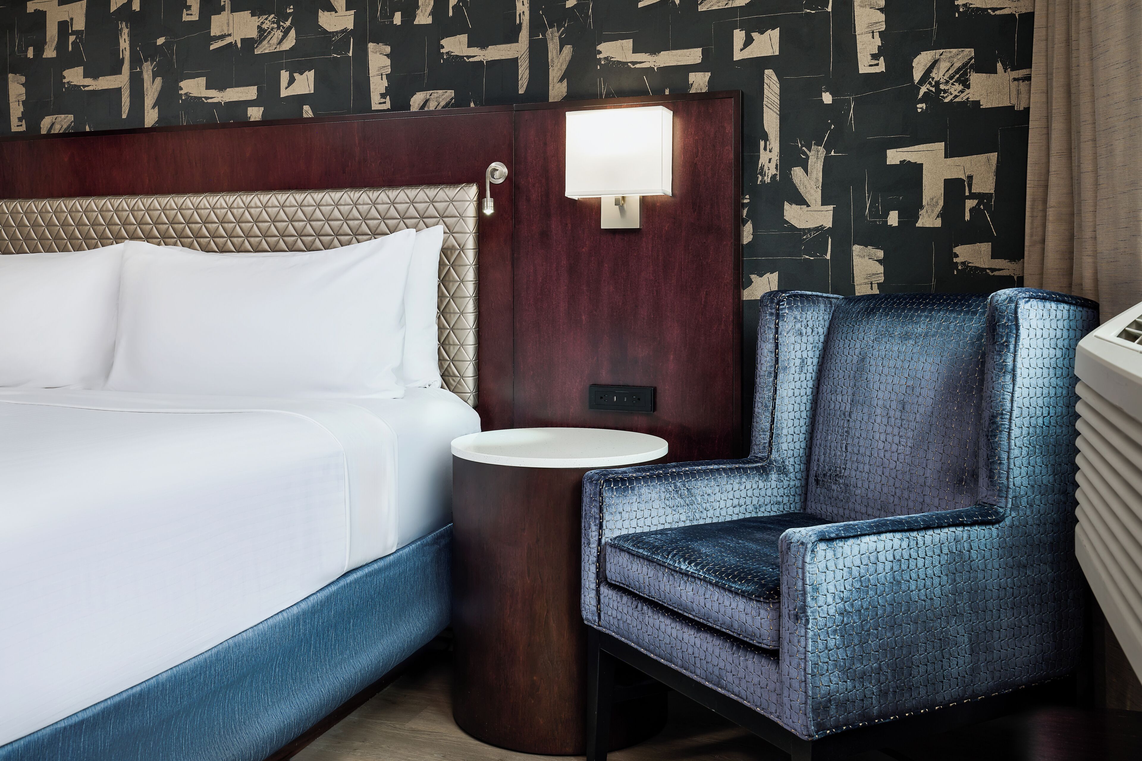 Une chambre d’hôtel avec un lit, une table de chevet ronde, une chaise et un mur orné d’un motif géométrique.