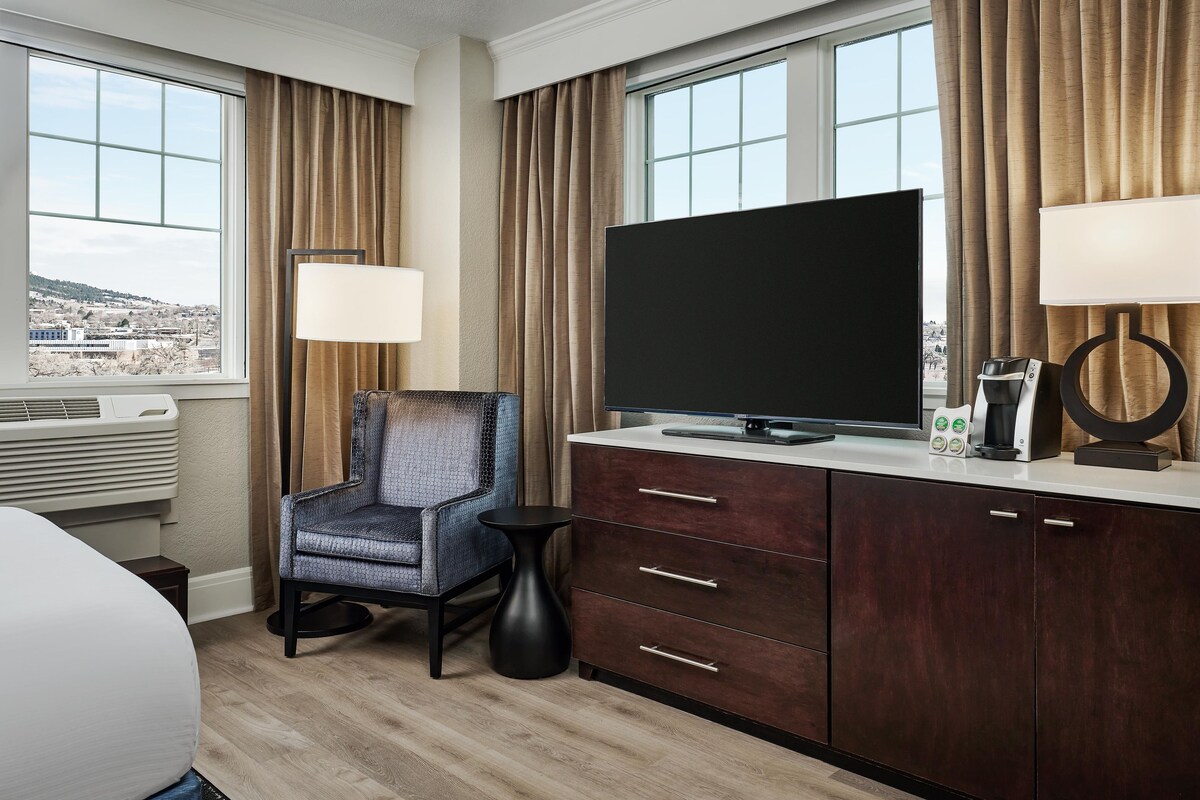 Quarto de hotel com televisão de tela grande, uma cadeira, mesa lateral pequena, armário com gavetas e janela com cortinas.