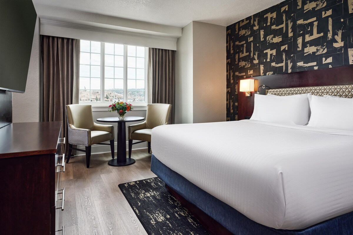 Quarto de hotel com uma cama grande, uma escrivaninha, duas cadeiras, uma pequena mesa com flores, vista pela janela e parede com padrão.