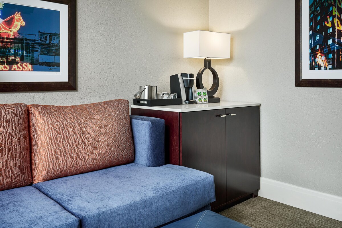 Quarto de hotel moderno com sofá, uma mesinha com luminária, cafeteira e obras de arte emolduradas nas paredes.