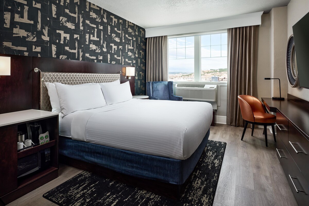 A hotel room with a large bed, a desk, a chair, a TV, and a window with a view.
