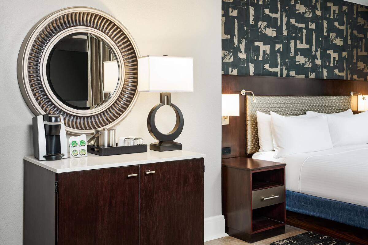 A hotel room with a large round mirror, a bedside table with a lamp, a bed with white linens, and a patterned wall.