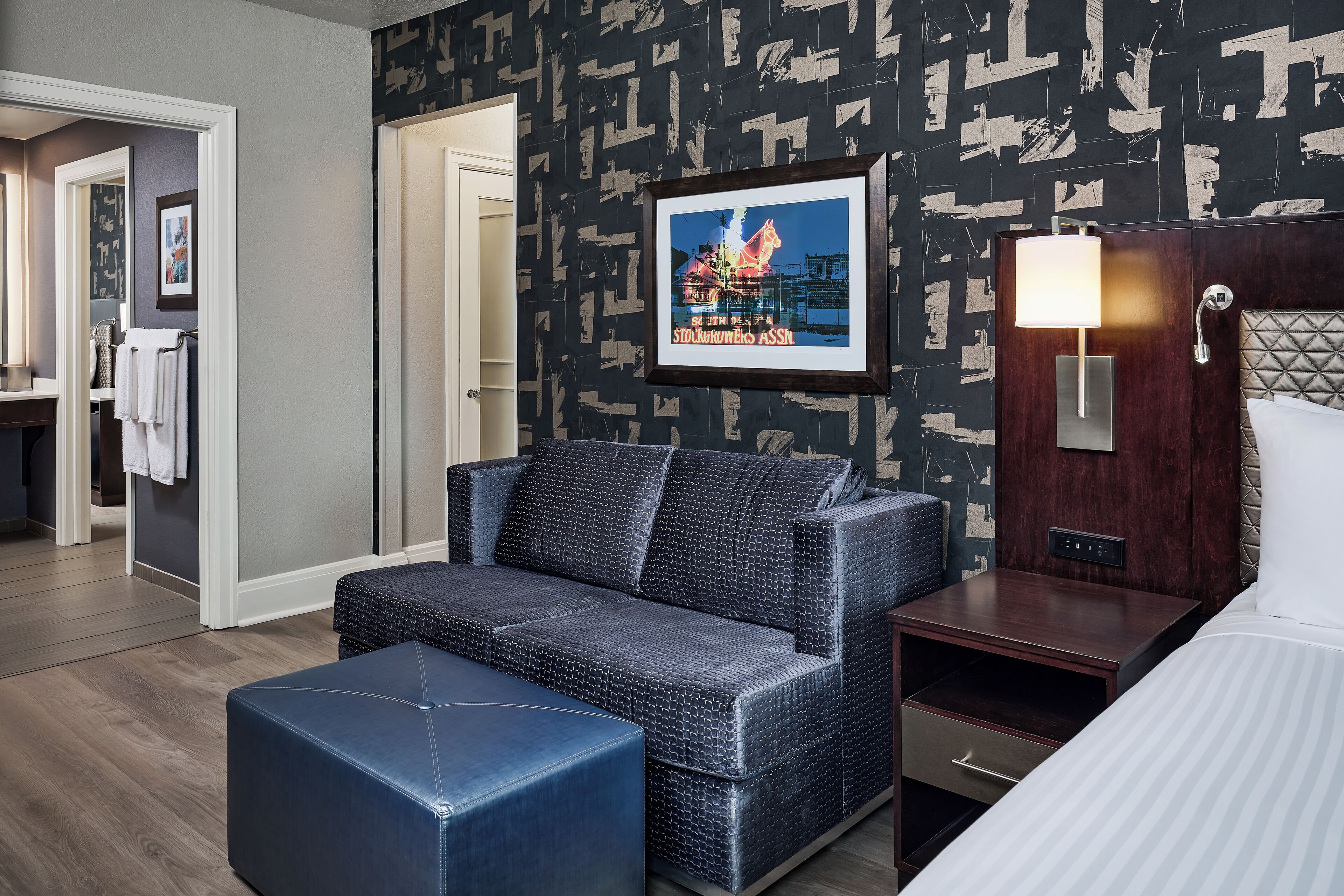 A hotel room with a sofa, ottoman, bedside table, lamp, and a framed picture on the wall.
