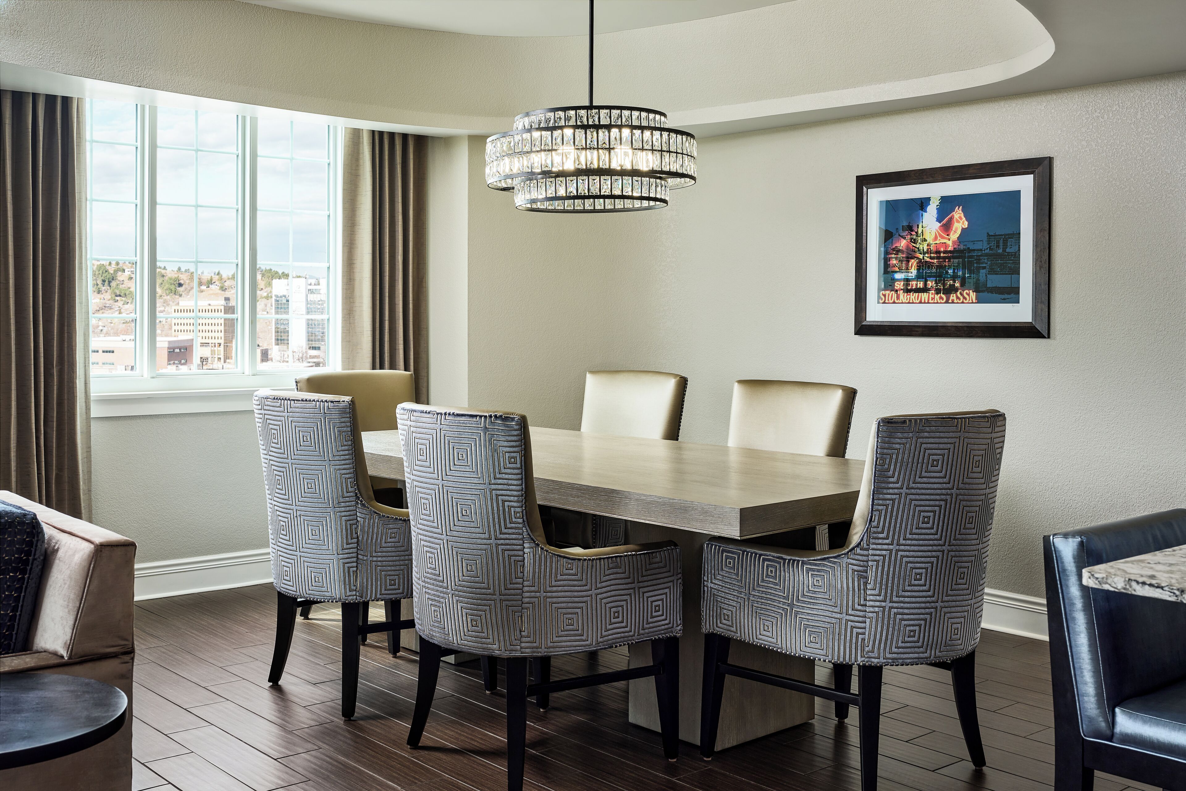 A dining area with a large table, four patterned chairs, a chandelier, a framed picture on the wall, and a window with curtains.
