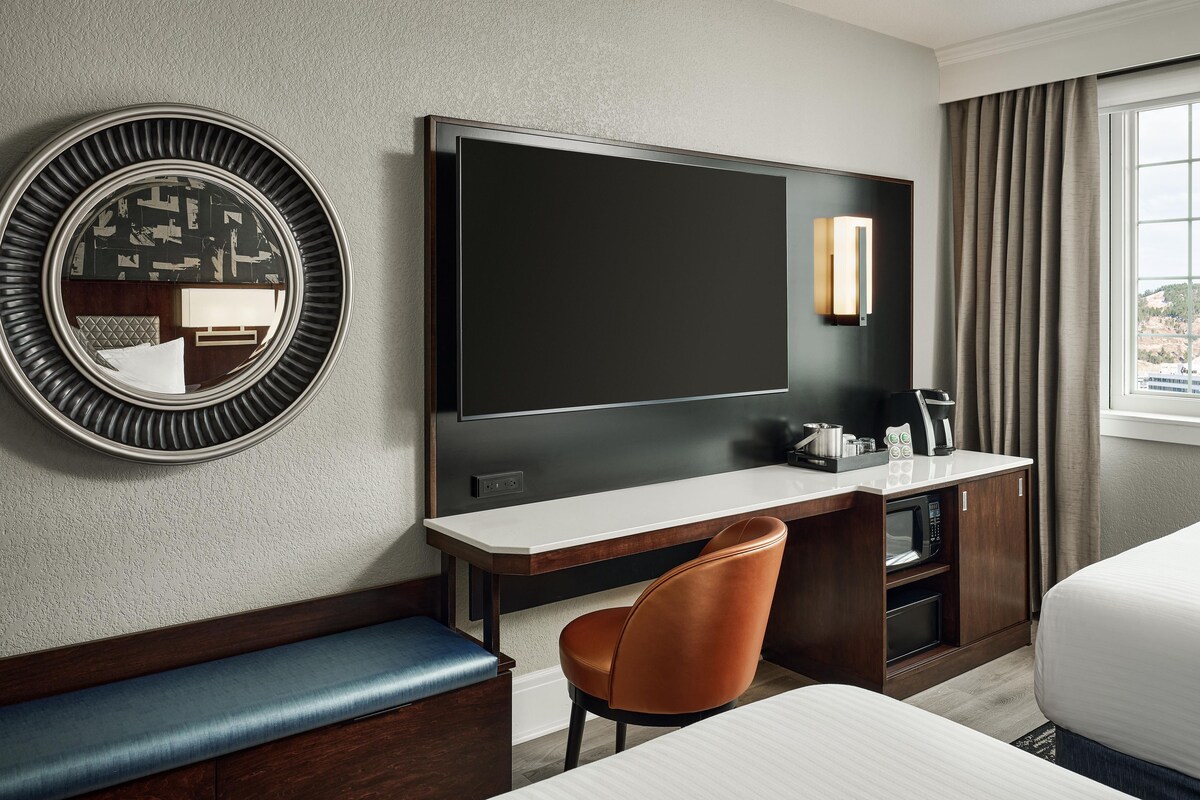Quarto de hotel com uma televisão de tela plana grande, um espelho redondo, uma escrivaninha de madeira, uma poltrona de couro marrom, uma cama e uma janela com cortinas.