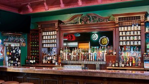 Un bar con mostrador de madera, taburetes y estantes llenos de botellas y vasos.