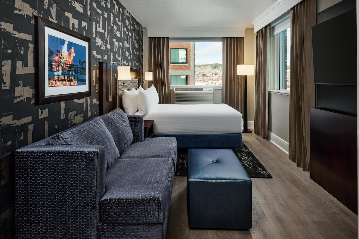 A hotel room with a large bed, a sofa, a small ottoman, a desk with a lamp, and a framed picture on the wall.