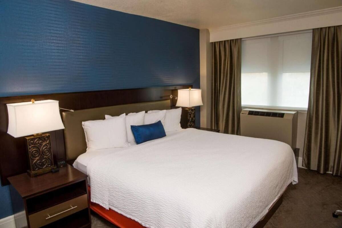 A hotel room with a large bed, two bedside tables with lamps, a window with curtains, and an air conditioning unit.