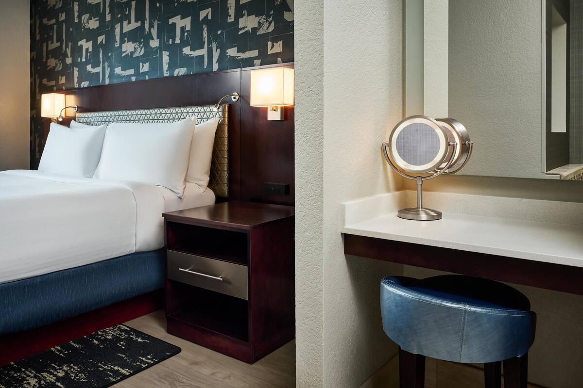 A hotel room with a bed, bedside table, lamp, mirror, and a desk with a chair.