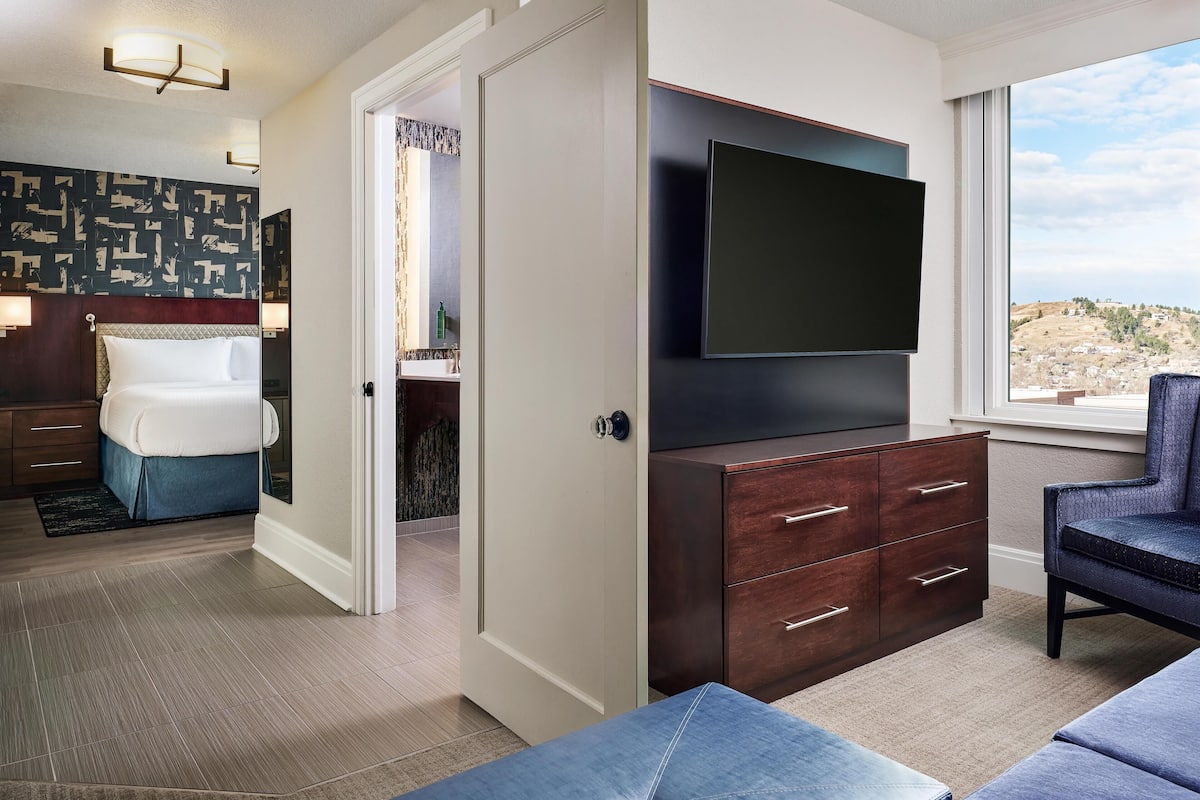 A modern hotel room with a large flat-screen TV, a comfortable bed, a seating area, and a view of the landscape.