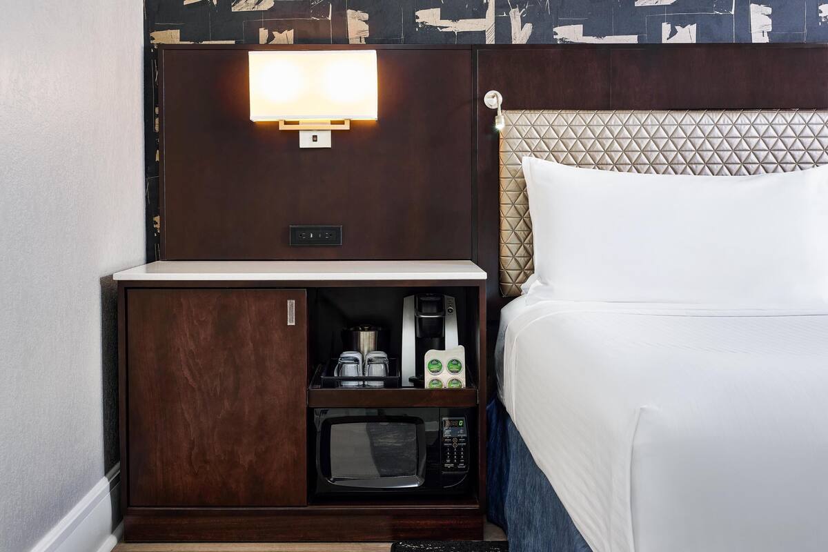 A hotel room with a bed, a bedside table with a microwave, a coffee maker, and a lamp.