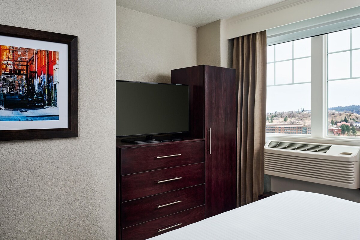 Quarto de hotel com uma cama, cômoda de madeira escura, televisão de tela plana e janela com vista para a cidade.