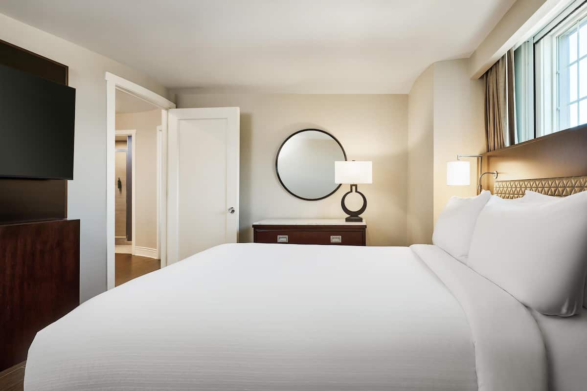 A hotel room with a large bed, a flat-screen TV, a round mirror, a bedside table with a lamp, and a view of the hallway.
