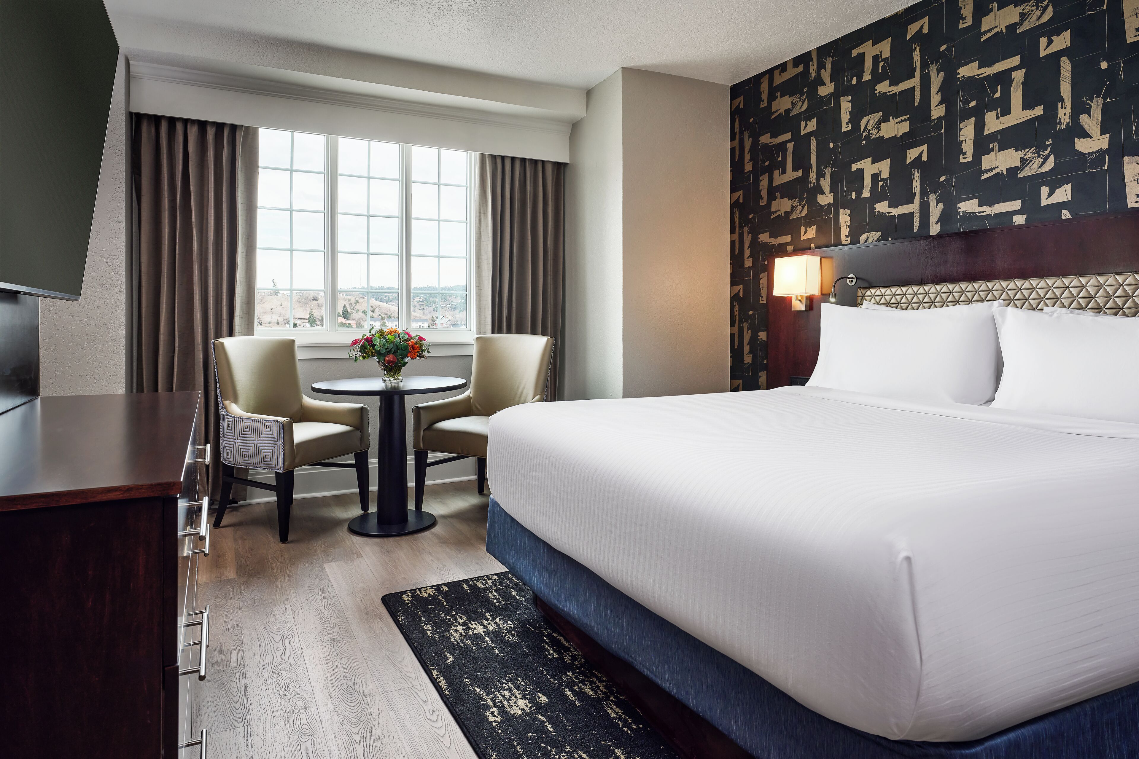 A hotel room with a bed, a desk, two chairs, a small table with flowers, a view through a window, and a patterned wall.