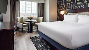 A hotel room with a bed, a desk, two chairs, a small table with flowers, a view through a window, and a patterned wall.