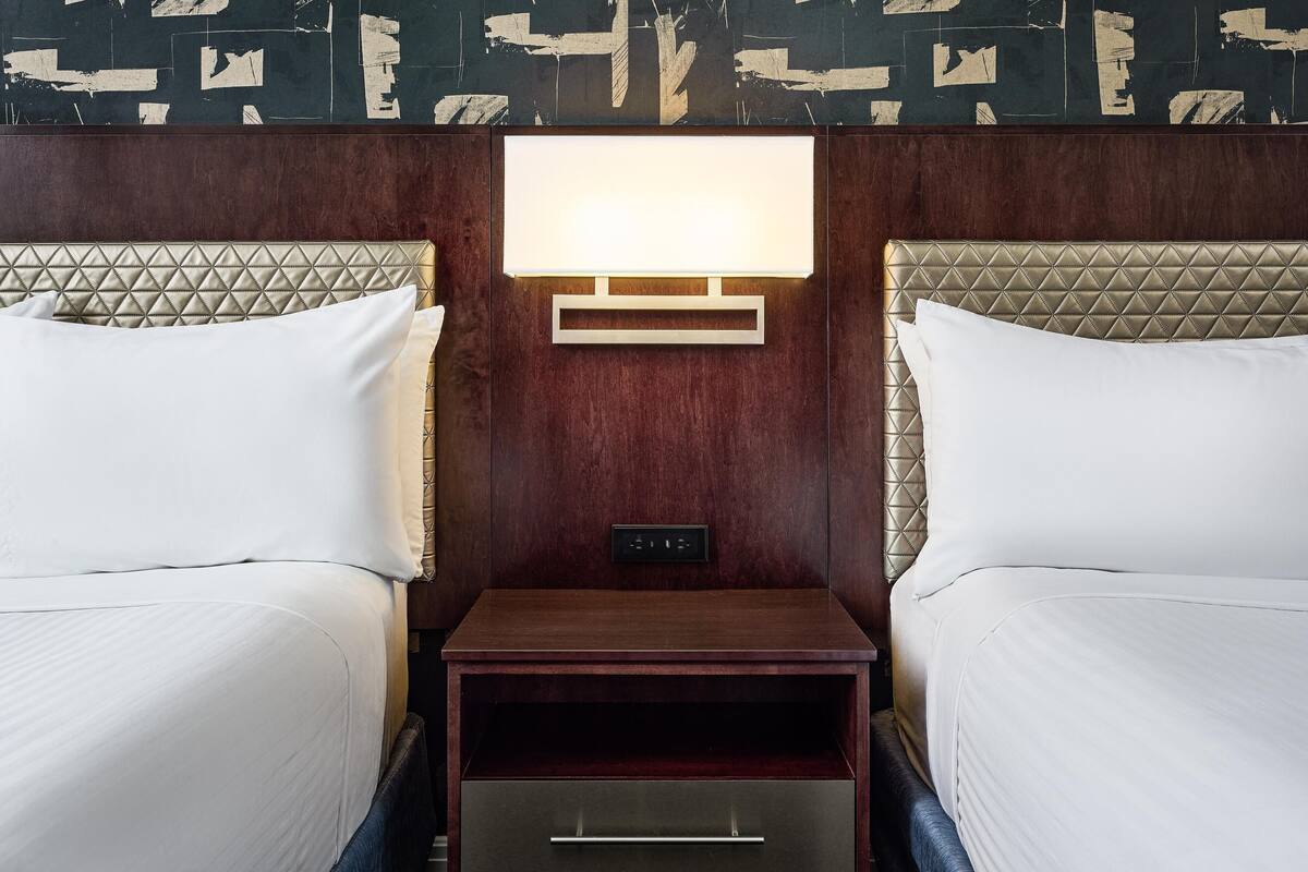 A hotel room with two beds, a nightstand with a lamp, and a wall-mounted control panel.
