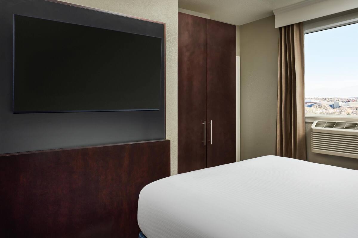 A hotel room with a bed, a flat-screen TV mounted on the wall, a wardrobe, and a window with curtains.