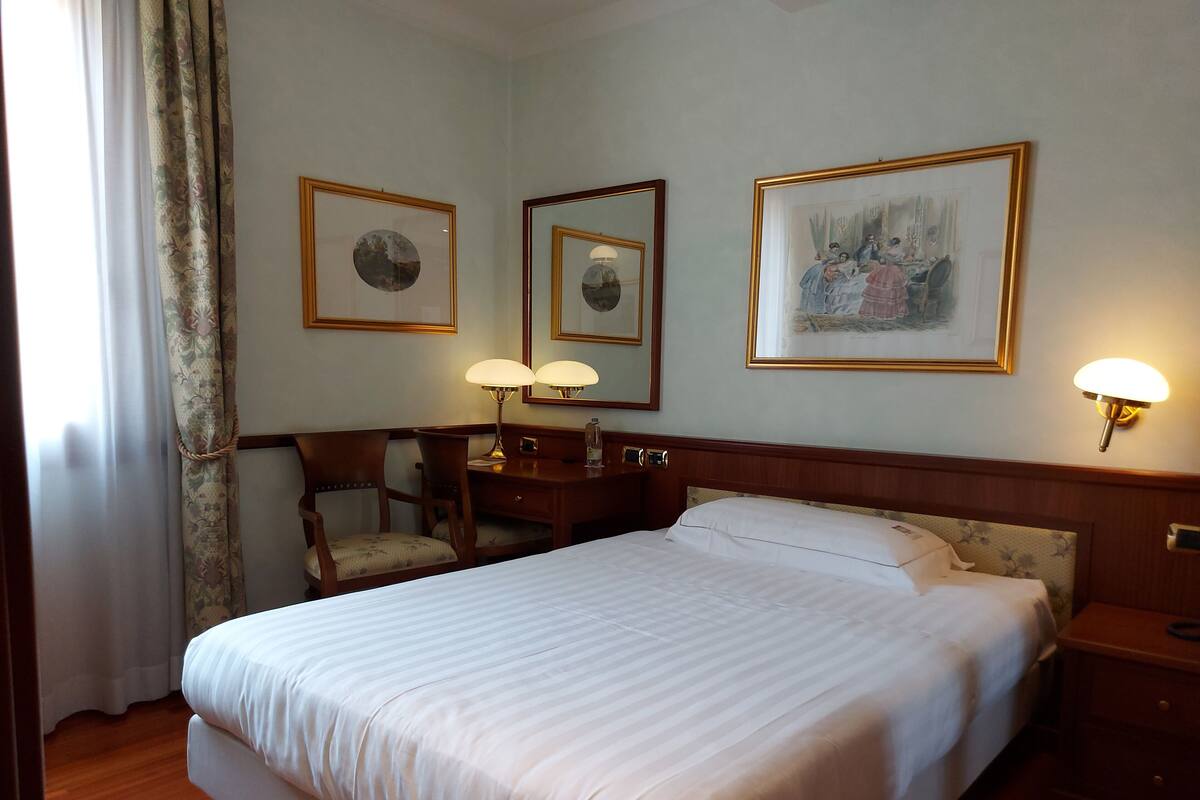 A hotel room with a large bed, a desk with a chair, two wall-mounted lamps, and framed artwork.