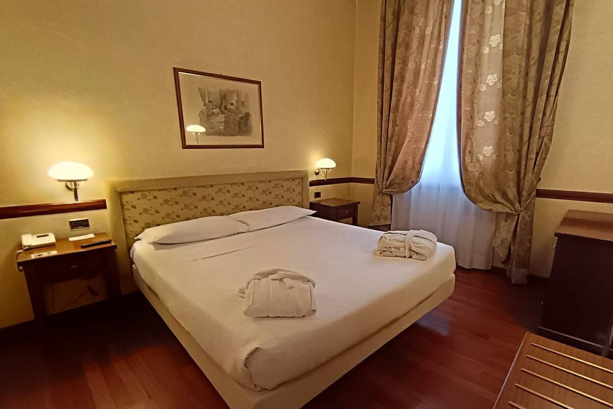 A hotel room with a bed, two bedside tables, a nightstand, a desk, a lamp, a framed picture on the wall, and a window with curtains.