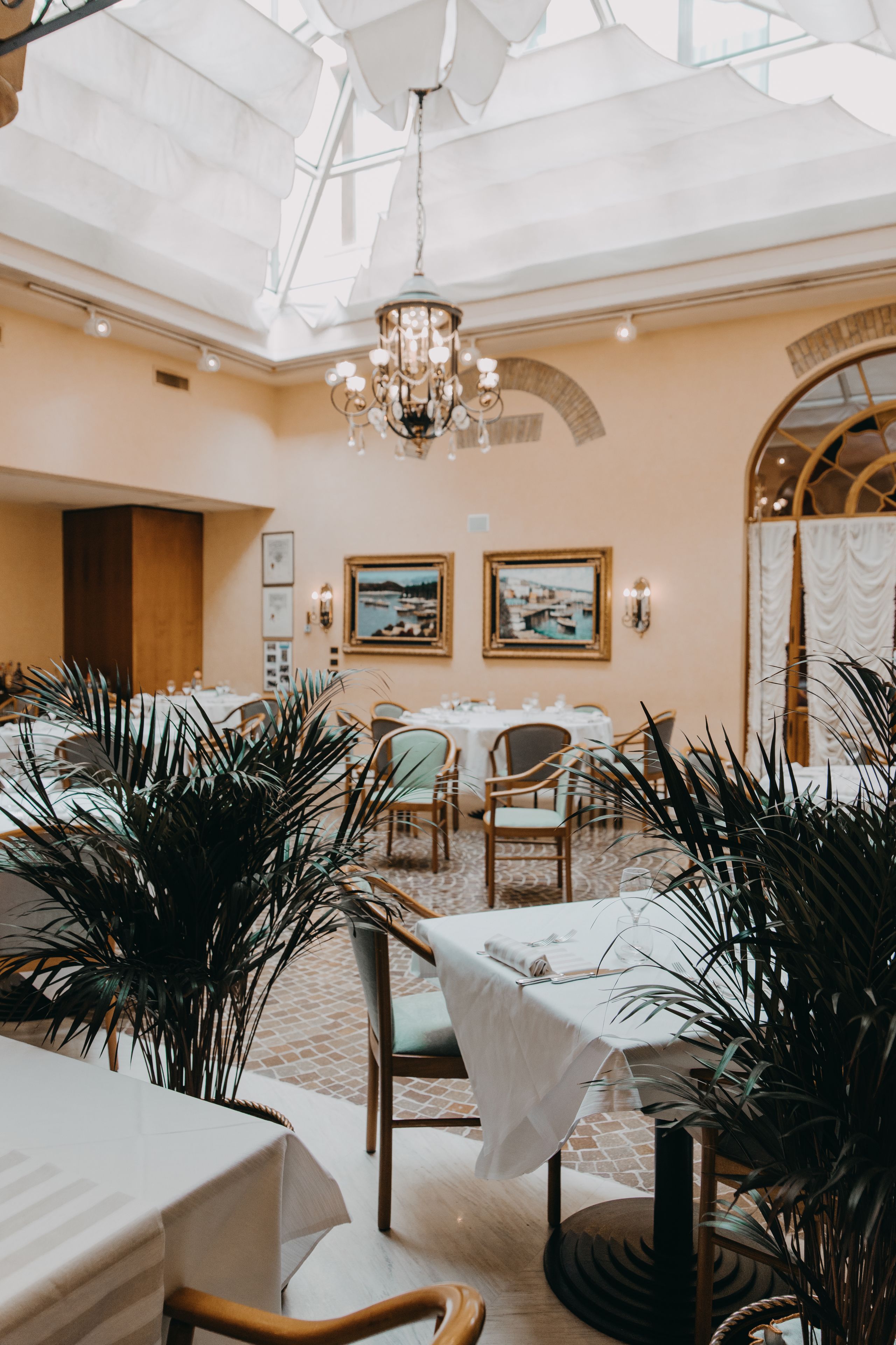 A dining area with tables set for guests, featuring a chandelier, framed artwork on the walls, and potted plants.