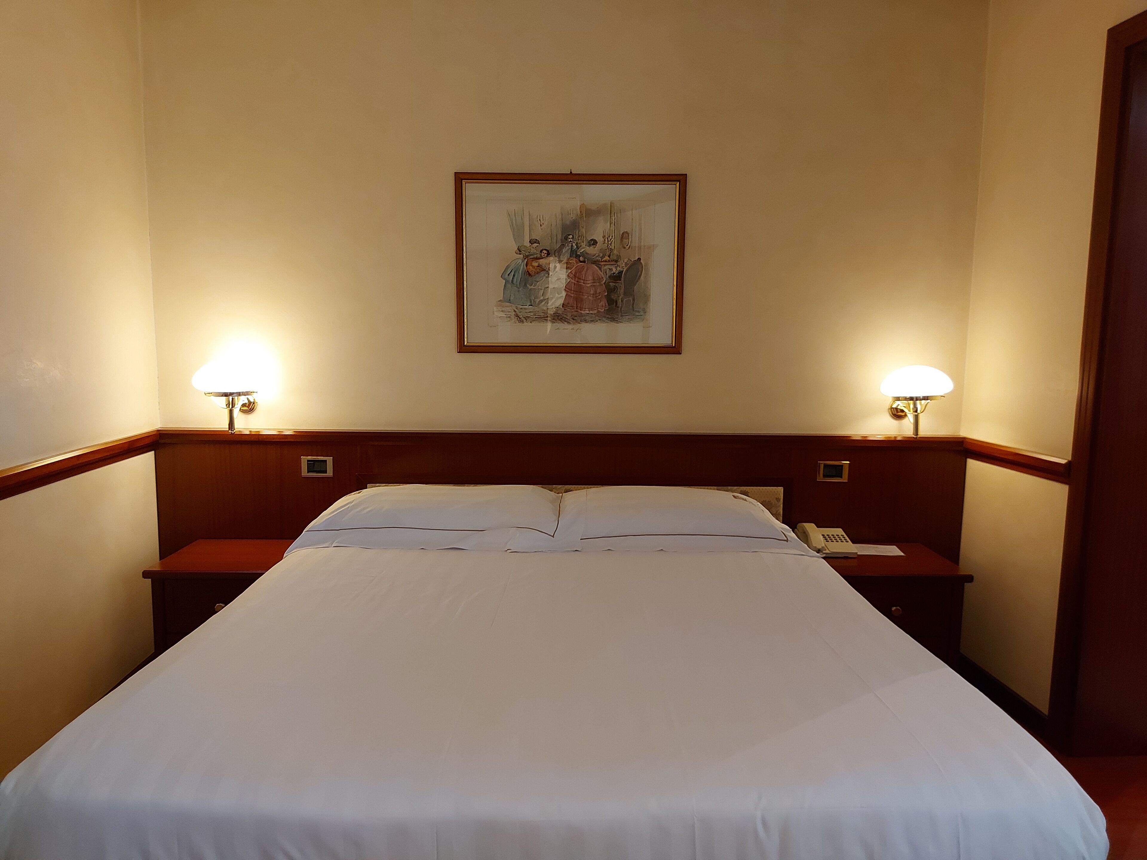A hotel room with a large bed, two bedside tables with lamps, a painting on the wall, and a telephone on one of the tables.