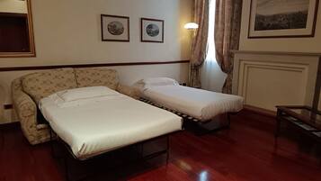 A room with a sofa and two single beds, wooden flooring, a standing lamp, and two framed pictures on the wall.