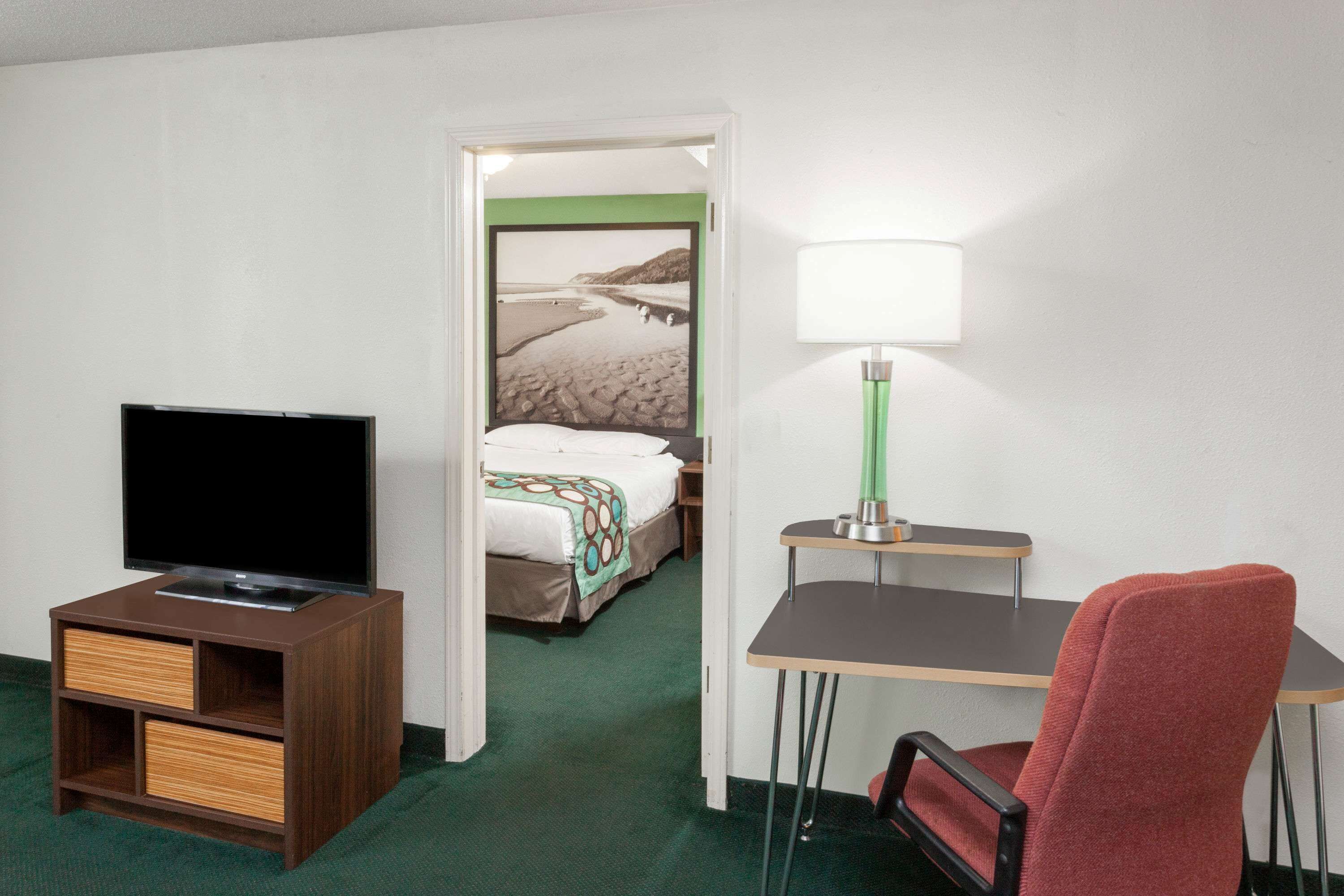 A hotel room with a television, a desk with a lamp, a red chair, and a bed with a green wall and a beach scene poster.