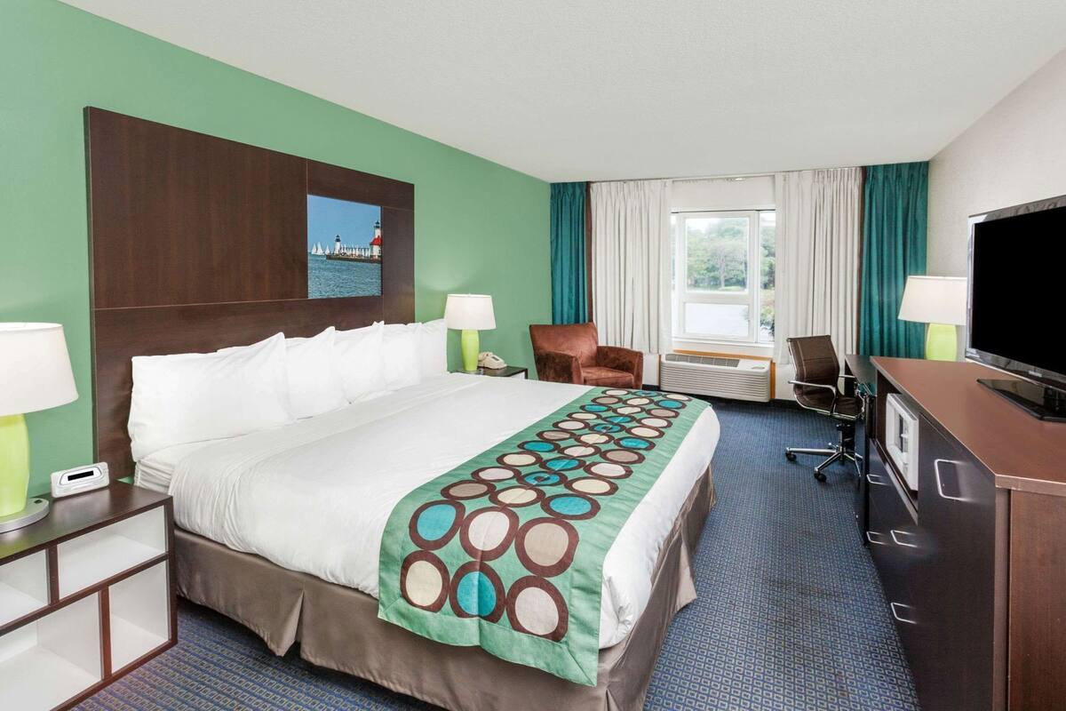 Una habitación de hotel con cama, escritorio, silla, televisor y ventana con cortinas.