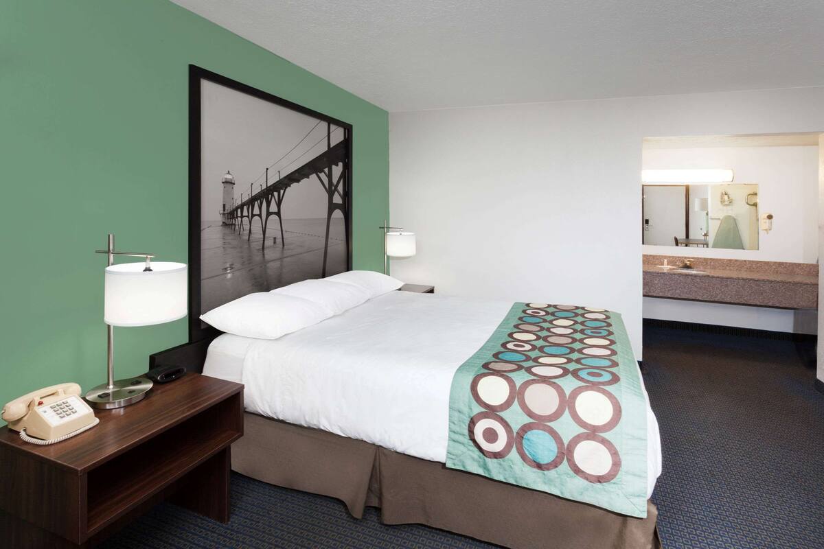 Una habitación de hotel con una cama, una mesita de noche con un teléfono, una lámpara de pared y un cuadro grande con un puente.