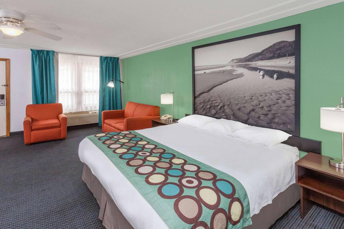 Habitación de hotel con una cama grande, dos sillones naranjas, un ventanal con cortinas verde azulado y un cuadro colgado en la pared de una playa, además de un ventilador de techo.