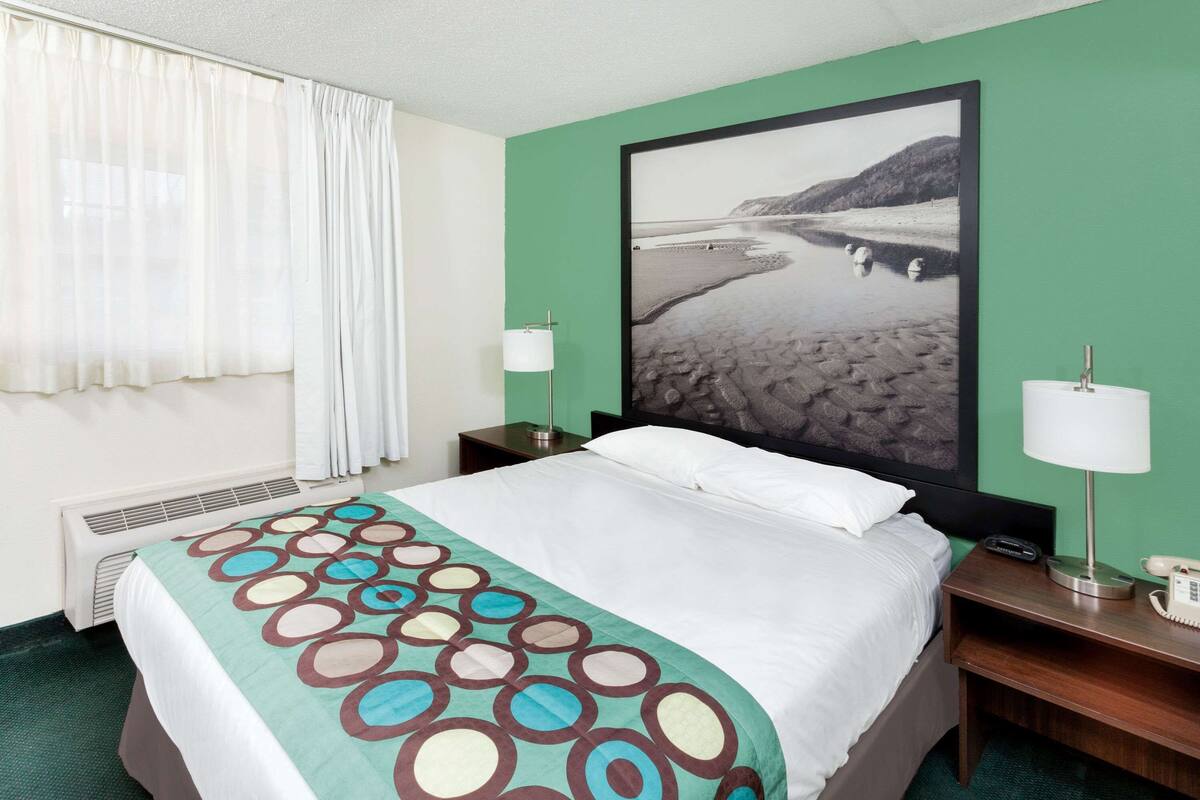 Habitación de hotel con cama, mesita de noche con lámpara, un cuadro grande enmarcado de una playa y una ventana con cortinas.