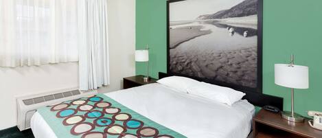 Quarto de hotel com cama, mesa de cabeceira com abajur, uma grande moldura com fotografia de uma praia e uma janela com cortinas.