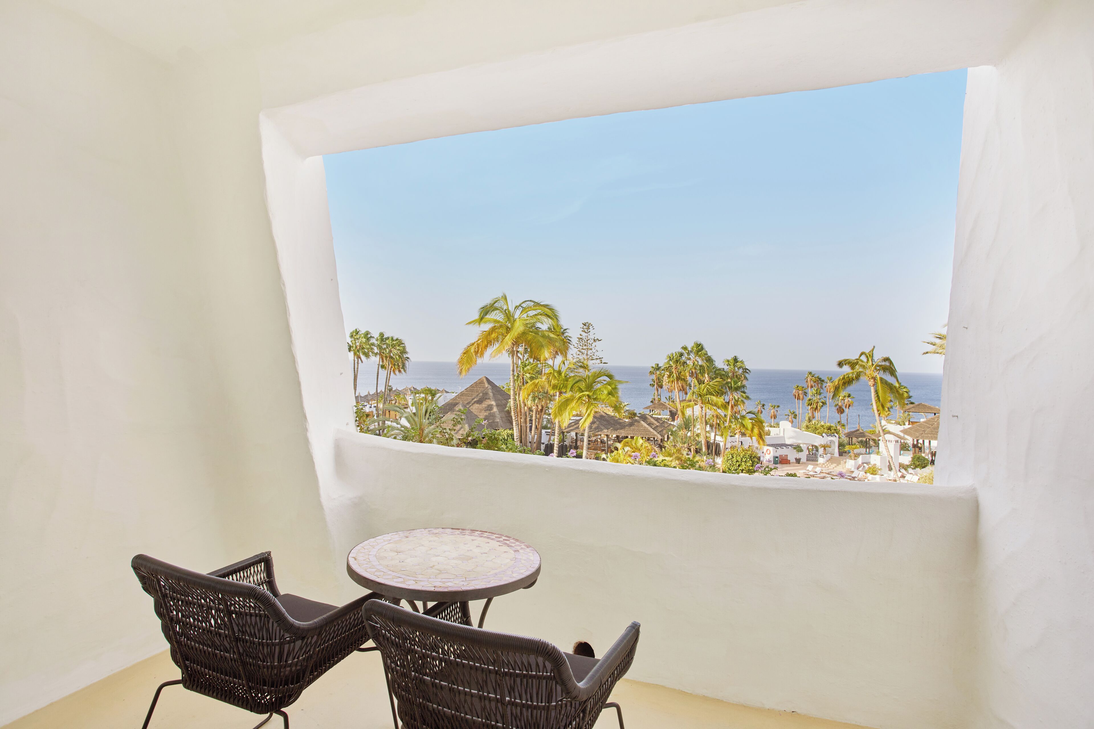 A balcony with wicker chairs and a small table, overlooking a beach with palm trees and a clear sky.