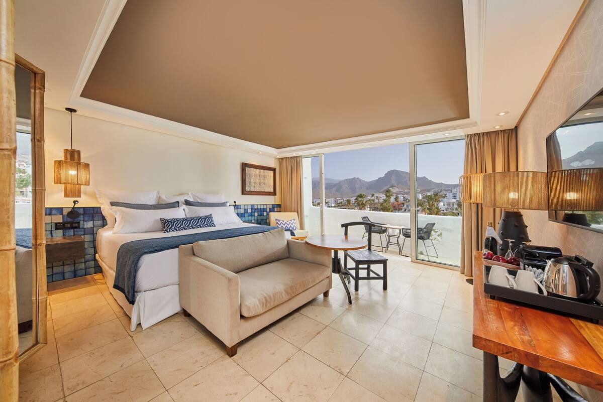 A hotel room with a bed, a sofa, a small table, and a balcony with a view of mountains.