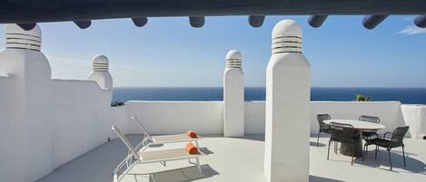 Una terraza en la azotea con paredes blancas, una pérgola, una mesa con sillas y vistas al mar.