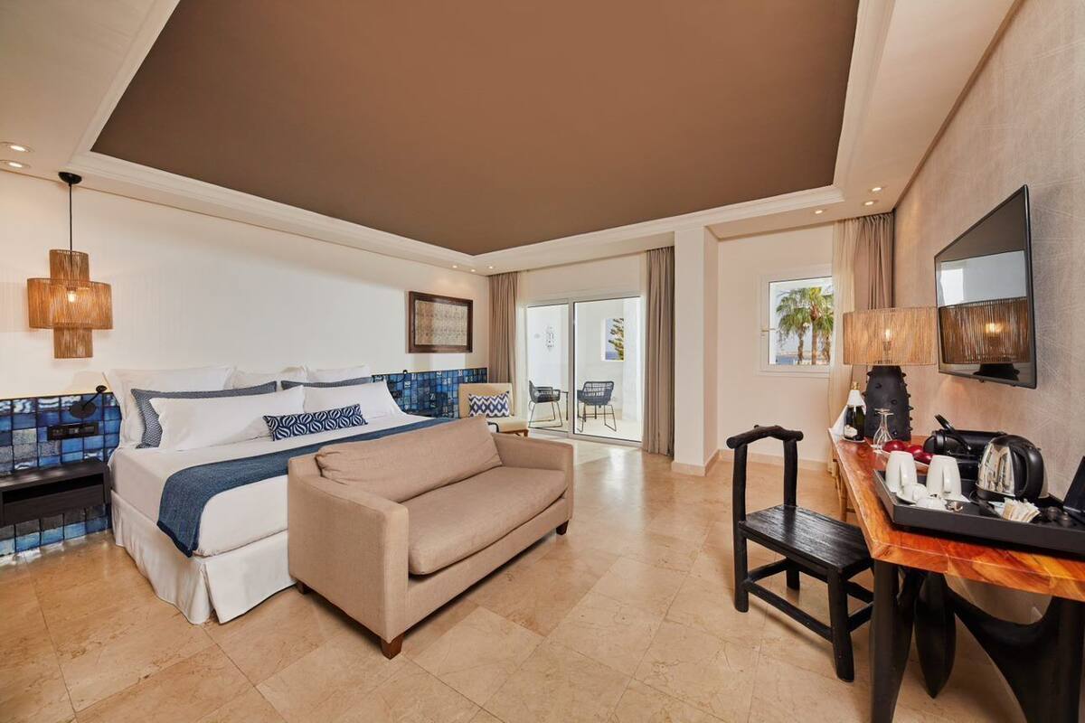 A hotel room with a bed, a sofa, a dining table with chairs, a TV, and a balcony with a view.