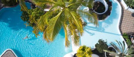 An aerial view of a resort with a large swimming pool surrounded by palm trees and a circular water feature.