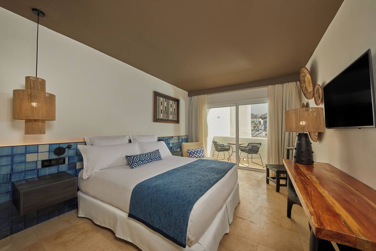 A hotel room with a bed, a nightstand, a wooden table, a TV, and a balcony with a view.