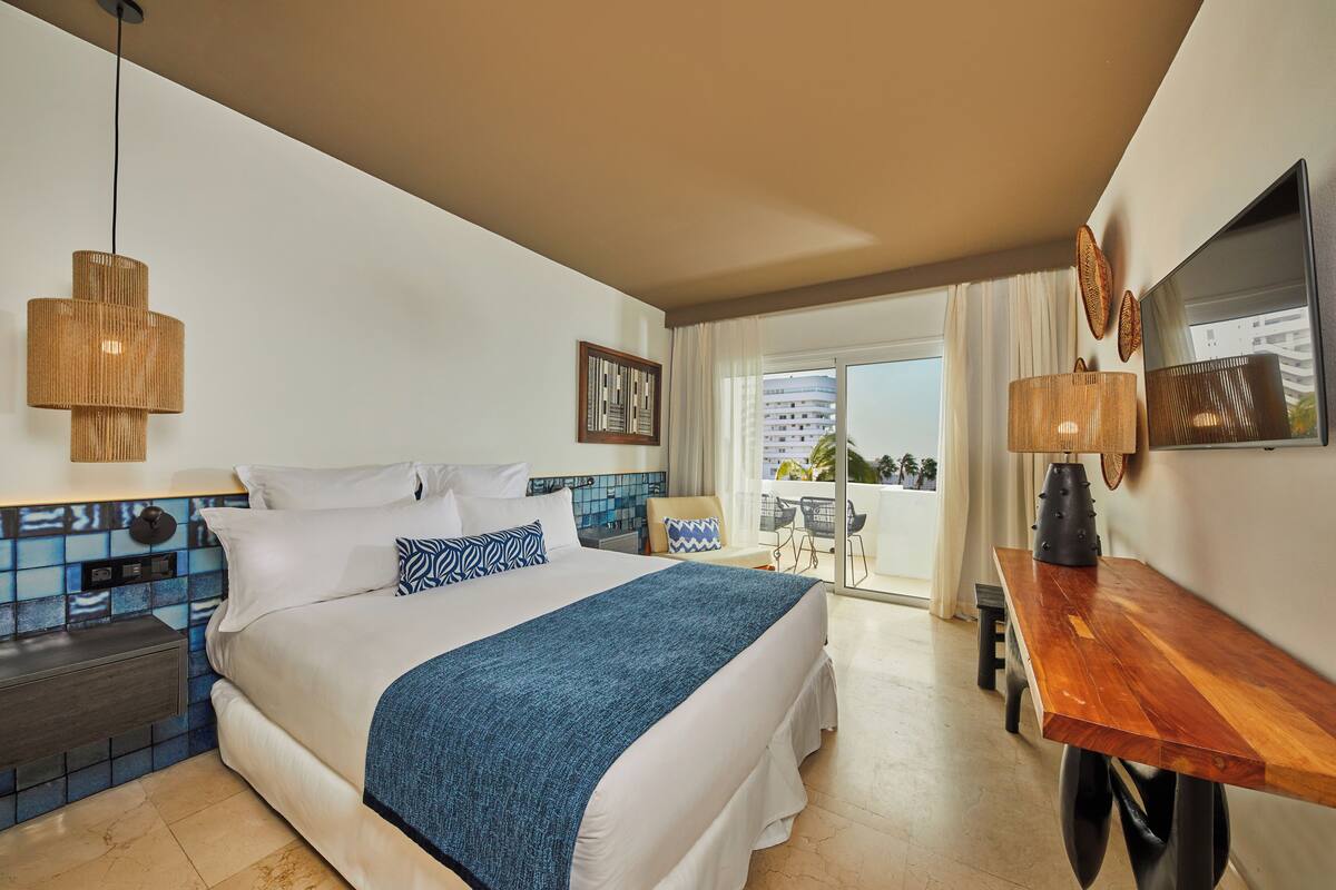 A hotel room with a large bed, a wooden desk, a balcony with a view, and a tiled wall.