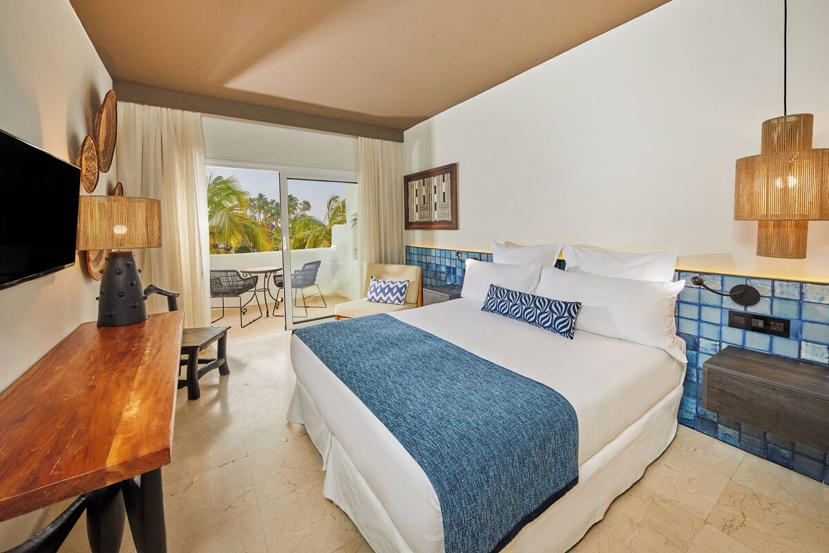 A hotel room with a large bed, a desk, a chair, a TV, and a balcony with a view of palm trees.