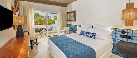 A hotel room with a large bed, a desk, a chair, a TV, and a balcony with a view of palm trees.