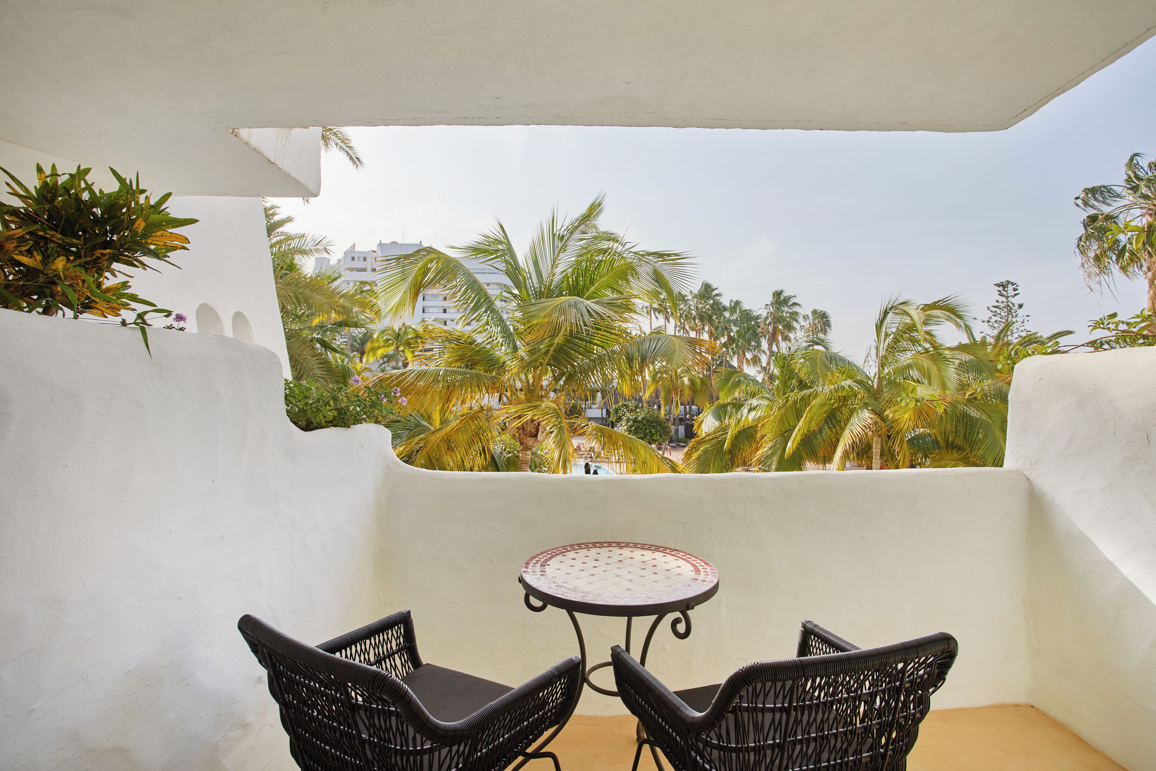 A balcony with a round table and two chairs, overlooking a garden with palm trees and a building in the background.