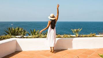 A woman in a white dress and hat standing on a tiled terrace with a view of the sea.