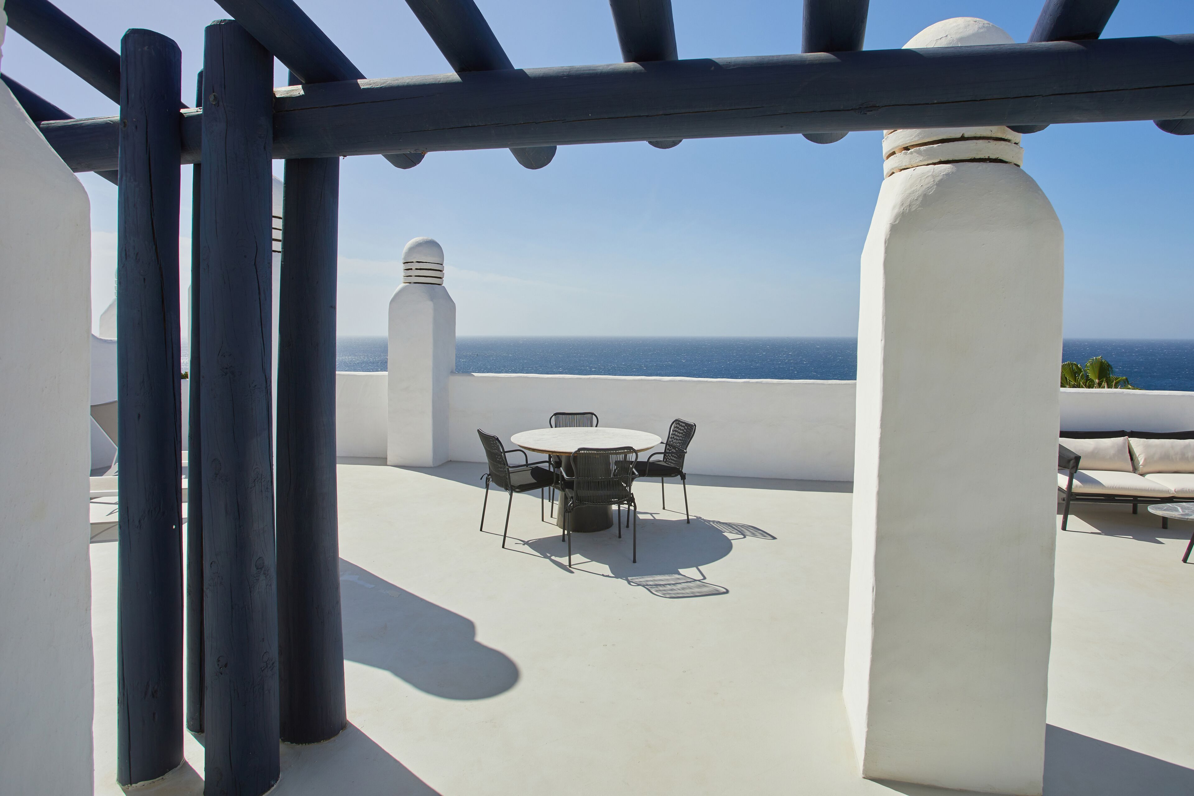 Una terraza en la azotea con una mesa redonda y sillas, un pilar blanco y vistas al mar.