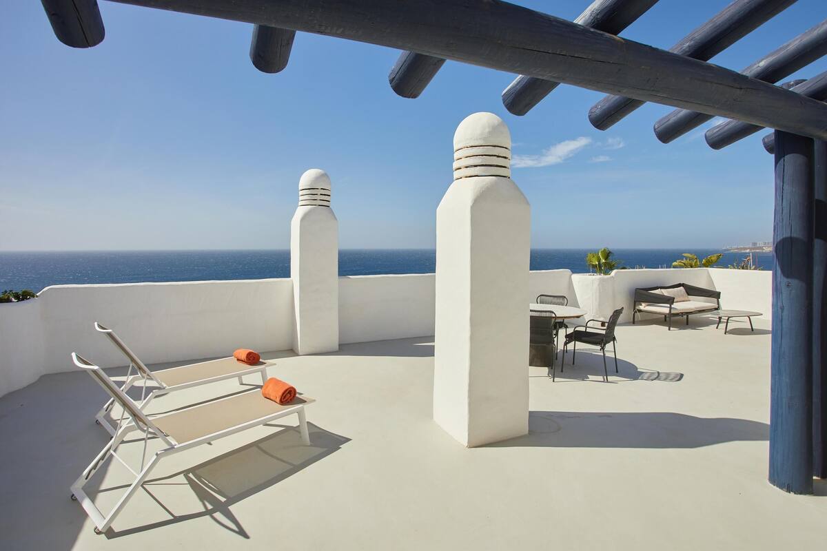 A rooftop terrace with a clear view of the sea, white pillars, and outdoor seating.