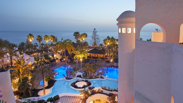 Zona de piscina en un resort con varias piscinas, palmeras y vista al océano al anochecer.