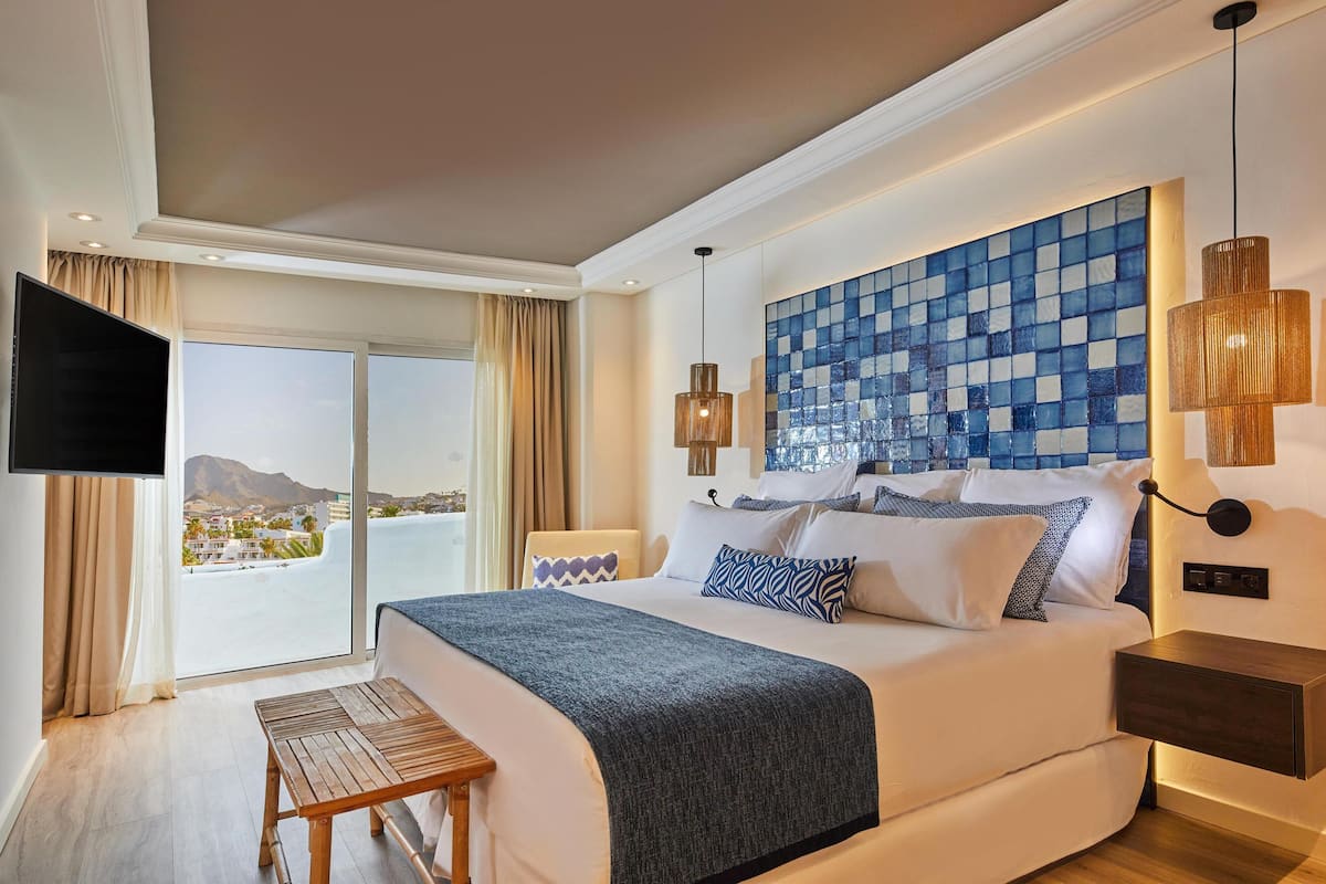 A hotel room with a large bed, a TV, a view of the sea, and a tiled wall.
