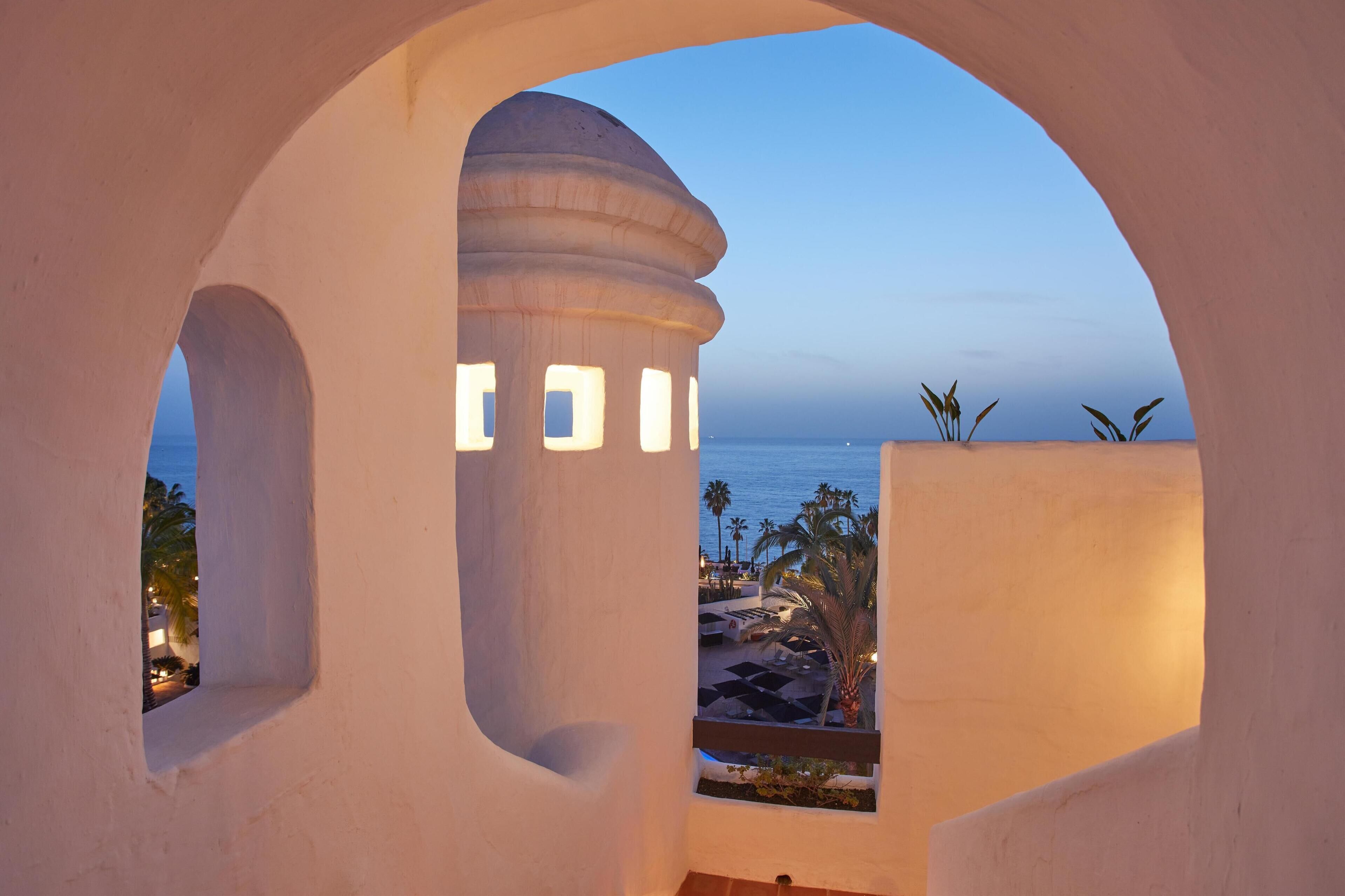 Vista a través de una ventana arqueada blanca, mostrando una zona de piscina y palmeras.